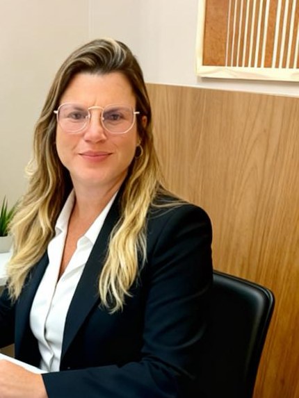 Luciane Barros sentada à mesa do consultório, sorrindo, com painel de madeira ao fundo.